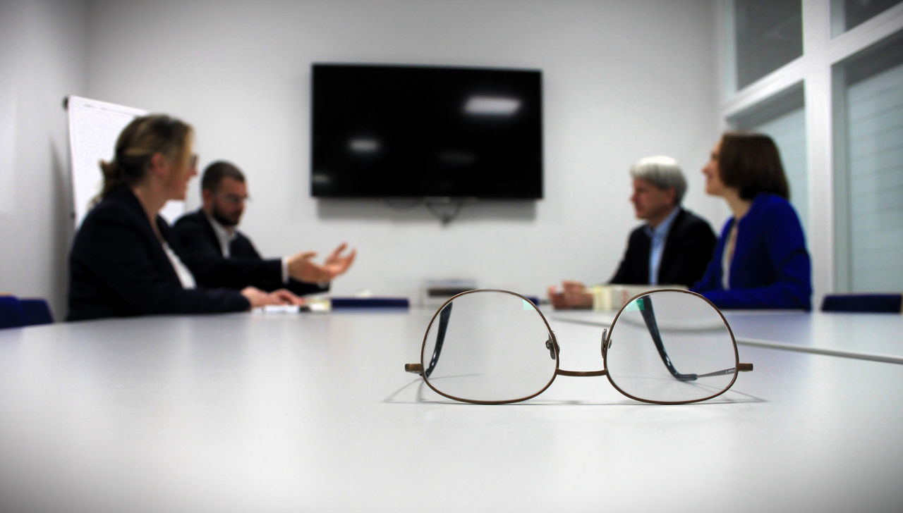 Ein Team von Kollegen berät über einen Gegenstand. Die im Vordergrund liegende Brille weist auf den Beratungscharakter hin.