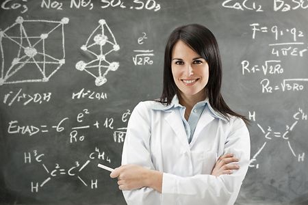 Eine l&auml;chelnde Wissenschaftlerin steht vor einer Tafel mit Formeln. 