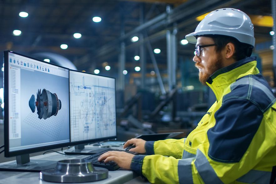 Mann in Arbeitskleidung in Industriehalle an PC mit Konstruktionszeichnungen.
