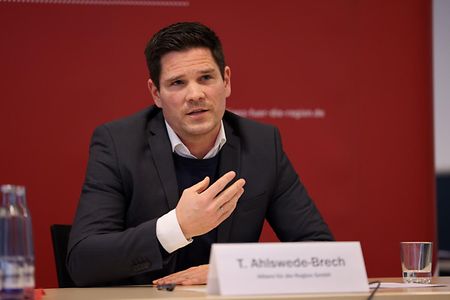 Thomas Alswede-Brech sitzt gestikulierend an einem Tisch und beantwortet Fragen. 