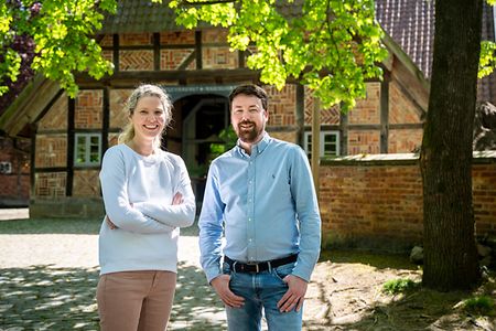 Inga Ali und Pascal Raschke stehen auf dem Hof des Biohotels WildLand, im Hintergrund das Fachwerkgeb&auml;ude.