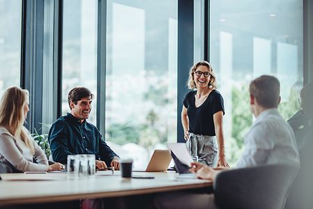 In einem Besprechungsraum mit Glasfront sind Kolleginnen und Kollegen lachend bei einem Meeting.