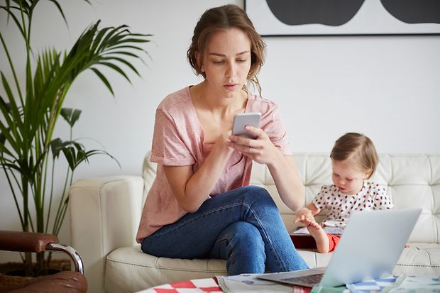 Eine junge Mutter recherchiert am Smartphone, während das Kind im Hintergrund spielt