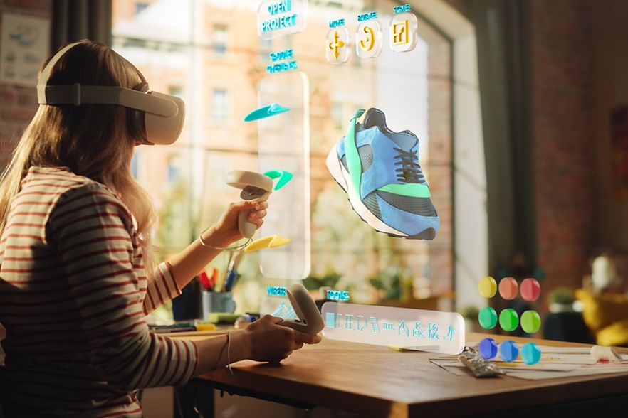 Frau sitzt am Tisch mit virtueller Brille, vor ihr "fliegt" z. B. ein Turnschuh