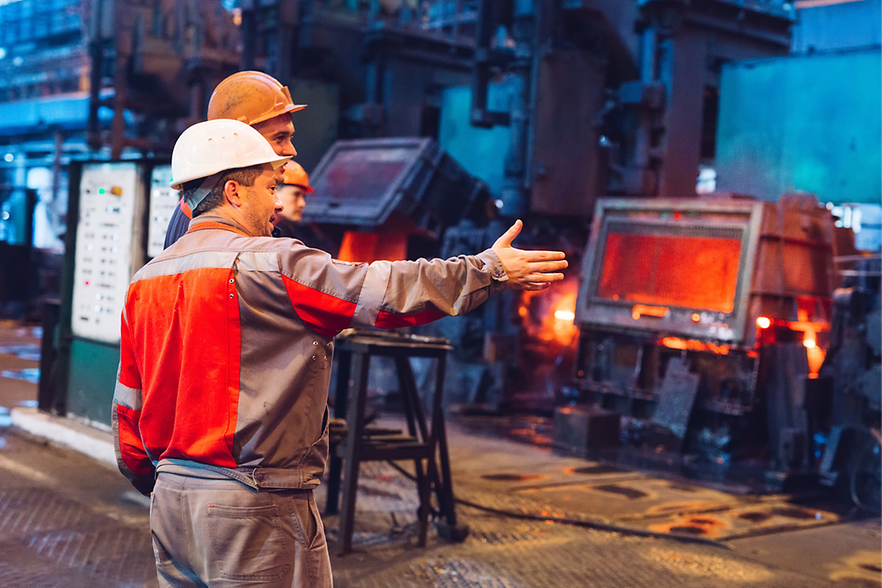 Konjunktur: Schwäche trifft auch Arbeitsmarkt drei Arbeiter in einem Stahlwerk stehen in der Produktionshalle. Einer zeigt auf den Ofen.