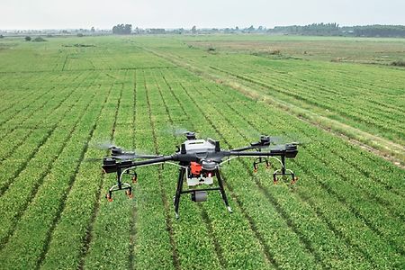 ein Feld, auf dem ein Traktor f&auml;hrt, dar&uuml;ber fliegt eine Drohne