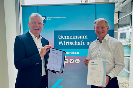 Zwei Männer stehen nebeneinander und lachen: Einer hält ein Tablet mit einem digitalen Zeugnis, der andere ein Papier-Zeugnis.