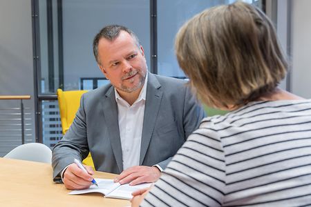 Ein Mann ber&auml;t eine Frau und zeigt mit einem Stift etwas in einem Heft, w&auml;hrend sie an einem Tisch sitzen