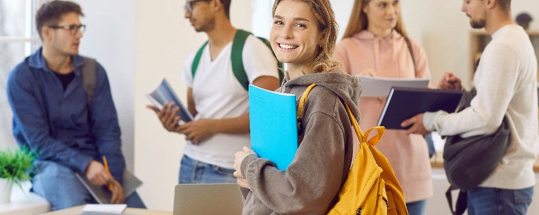 Ein l&auml;chelndes M&auml;dchen mit Rucksack und eine Gruppe im Hintergrund