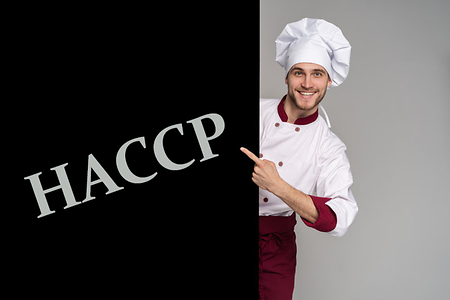 Ein Koch zeigt auf ein Schild, darauf steht HACCP (Hazard Analysis and Critical Control Points)