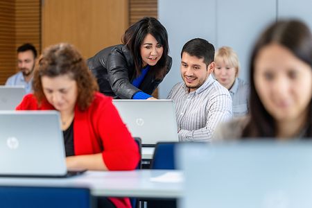 Teilnehmer einer Schulung arbeiten an ihren Laptops. Eine Dozentin steht neben einem Teilnehmer und zeigt auf seinen Bildschirm.