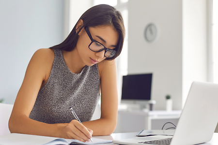 Junge Frau, die an ihrem Tisch am Laptop sitzt, neben dem B&uuml;cher und ein Kaffee stehen
