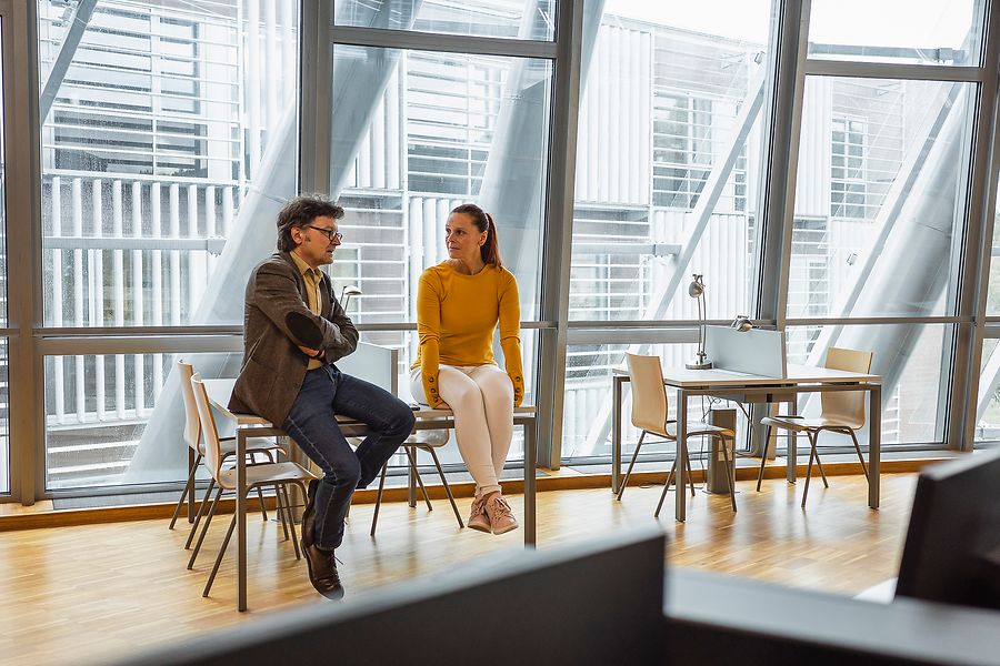 Älterer Mann und mittelalte Frau unterhalten sich im Büro und sitzen beide auf dem Tisch
