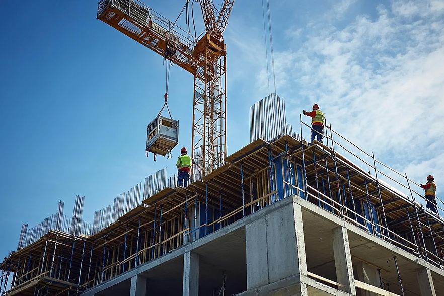 Ein Kran, der einen schweren Block auf eine Baustelle hebt. Auf dem Geb&auml;ude stehen drei Bauarbeiter.