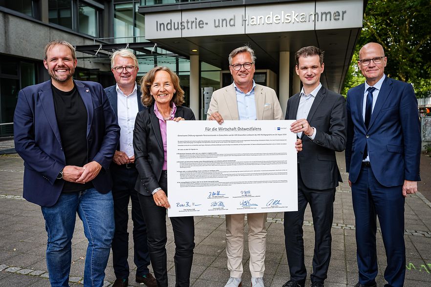 Sechs Personen stehen vor dem IHK-Geb&auml;ude in Bielefeld und halten ein gro&szlig;es Dokument zur Wirtschaft in Ostwestfalen.