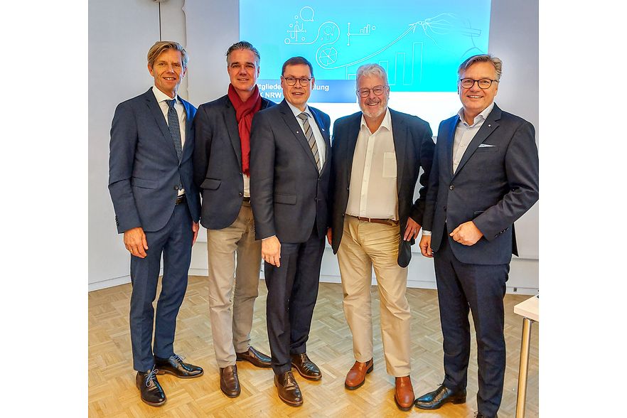 F&uuml;nf M&auml;nner im Anzug bei einer Veranstaltung, stehend vor einer Pr&auml;sentationsfolie.