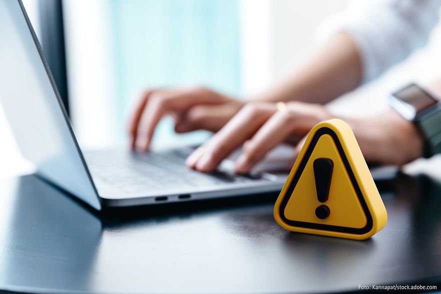 H&auml;nde tippen auf Laptop-Tastatur, im Vordergrund steht ein gelbes Warnsymbol mit Ausrufezeichen &ndash; Hinweis auf digitale Gefahr.