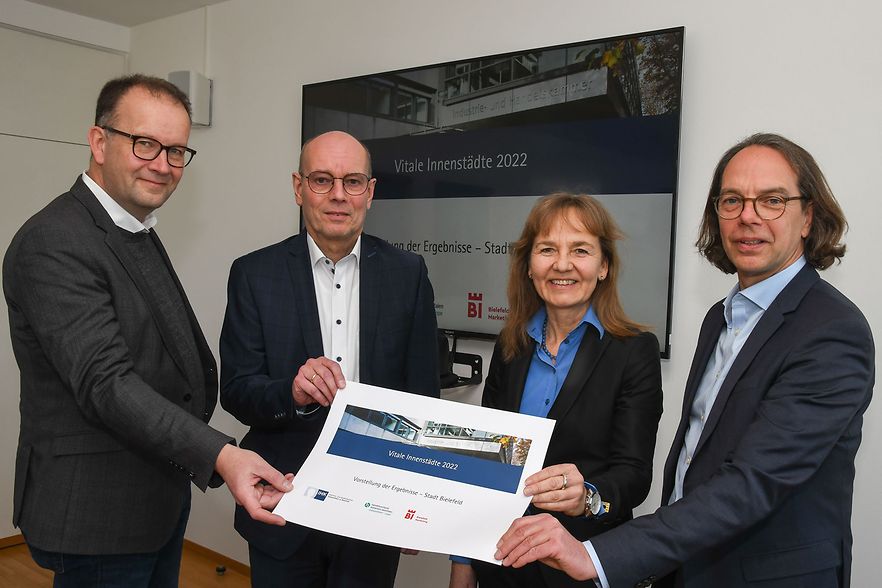 Vier Personen in Businesskleidung stehen vor einem Bildschirm mit der Aufschrift &bdquo;Vitale Innenst&auml;dte 2022&ldquo;.