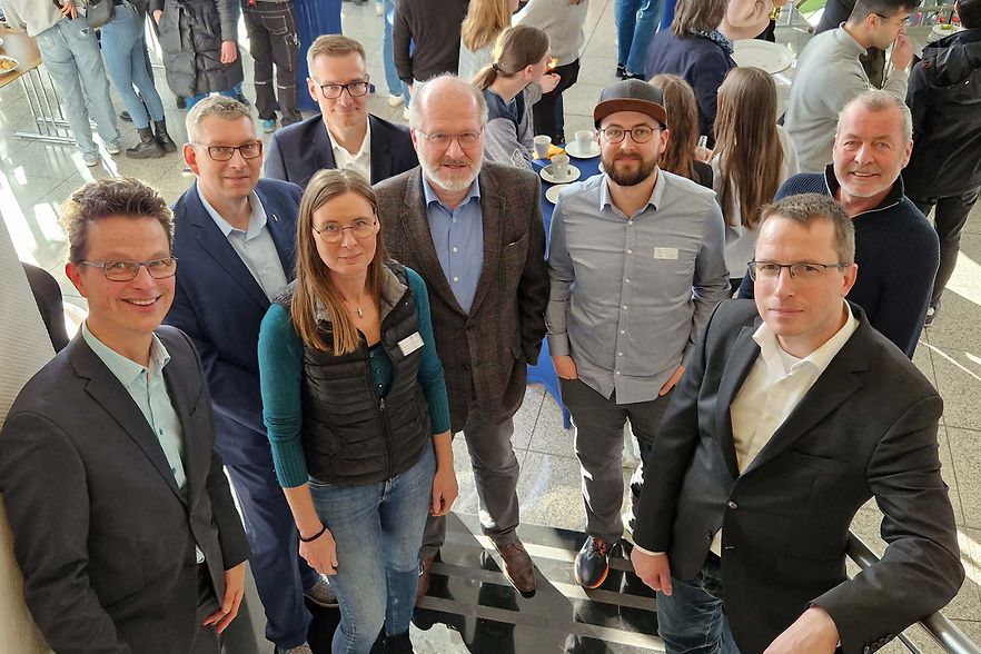 Gruppe von acht Personen, die l&auml;chelnd in die Kamera schauen, bei einer Auftaktveranstaltung in einem hellen Foyer.
