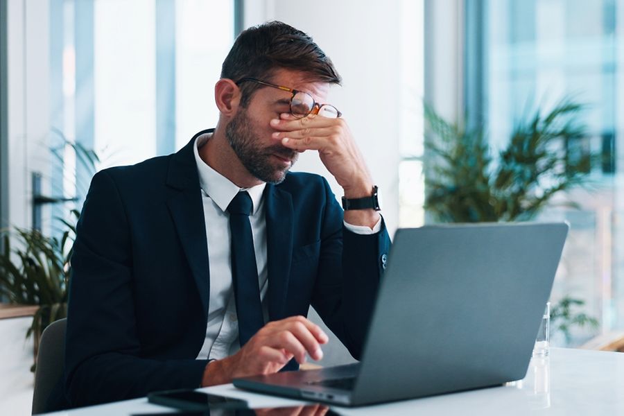 Mann im Anzug sitzt vor seinem Laptop und reibt sich verzweifelt die Augen