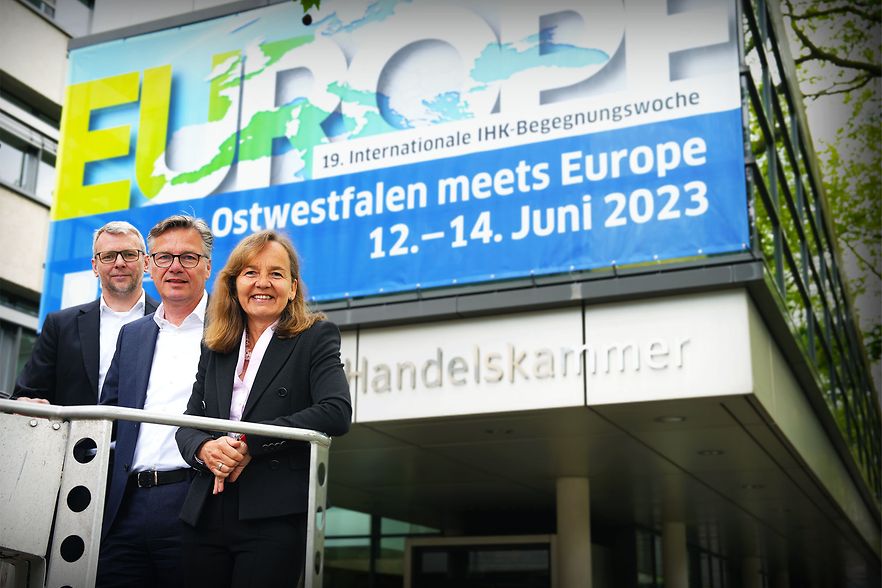 Drei Personen stehen vor der IHK mit einem Banner zur Veranstaltung &bdquo;Ostwestfalen meets Europe&ldquo; im Juni 2023.