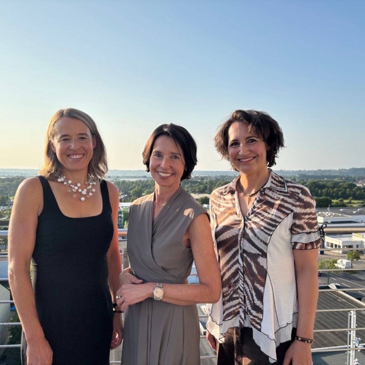Drei Frauen stehen im Sonnenschein auf einer Dachterrasse und lächeln in die Kamera. 