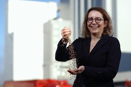 Person h&auml;lt Getreide in der Hand und l&auml;sst es vor gro&szlig;en Silos und roten LKWs durch die Finger rieseln.