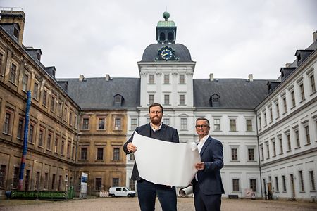 Zwei Personen halten gemeinsam einen ausgerollten Plan aus Papier und stehen vor H&auml;usern