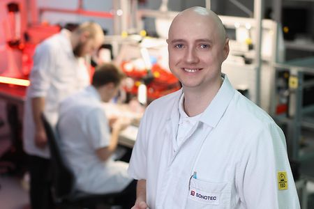 Person in wei&szlig;em Laborkittel h&auml;lt eine Leiterplatte, im Hintergrund arbeiten weitere Personen an technischen Ger&auml;ten.