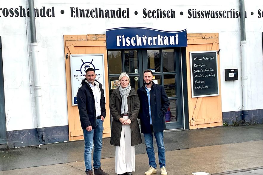 Zwei M&auml;nner und eine Frau stehen vor einer Eingangst&uuml;r mit der Aufschrift Fischverkauf.