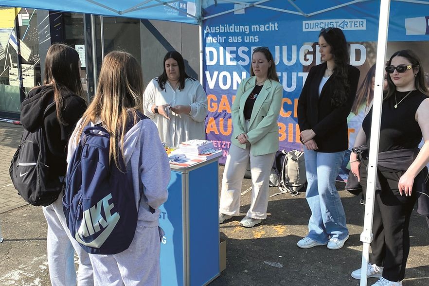 4 junge Frauen stehen an einem Messestand, davor sind 2 Sch&uuml;lerinnen