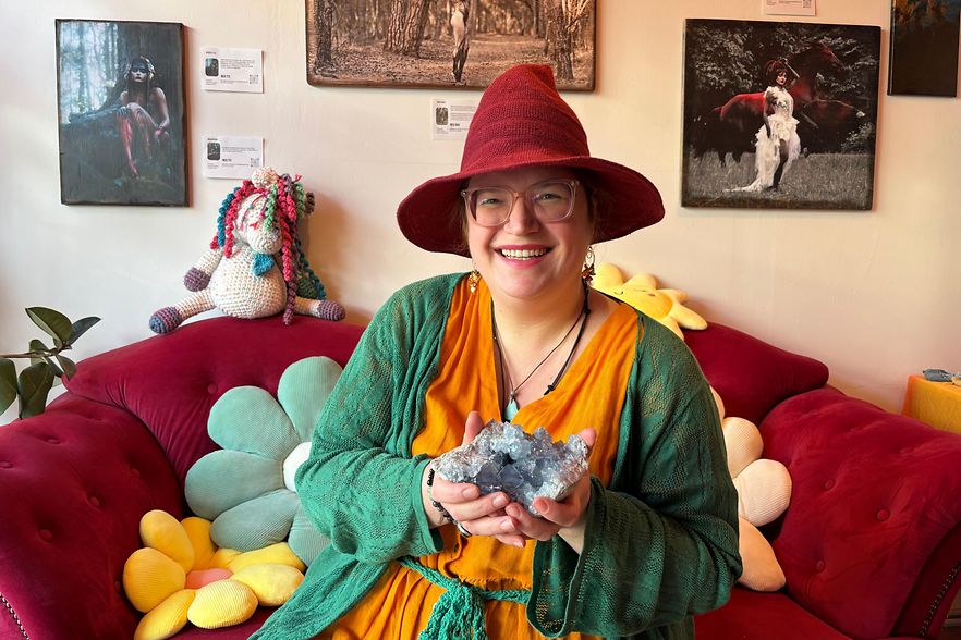 Eine bunt gekleidete Frau mit rotem Hut sitzt auf einem roten Sofa und h&auml;lt Kristalle in der Hand.