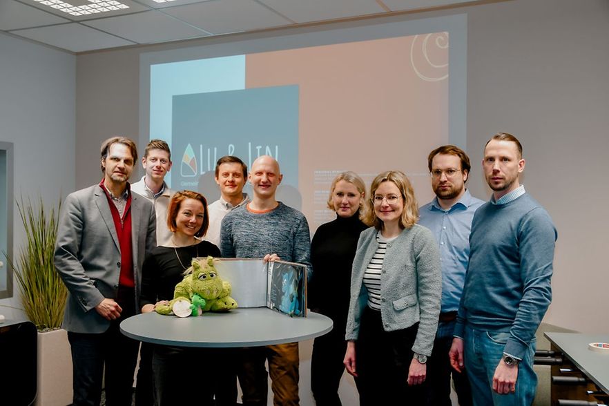 Gruppe vor Pr&auml;sentationsfolie mit Tisch und Maskottchen."