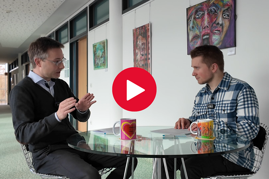 Zwei Personen sitzen an einem Tisch und unterhalten sich. In der Mitte des Bildes ist ein Play-Button zu sehen. 