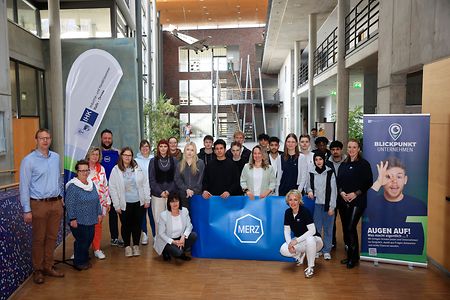 Gruppenfoto mit MERZ‑Banner in einem modernen Geb&auml;ude.