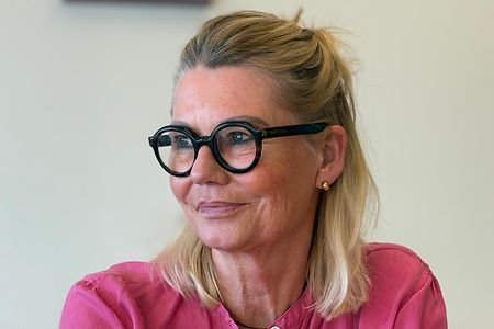 Eine Frau mit Brille und rosa Bluse sitzt und schaut zur Seite. 