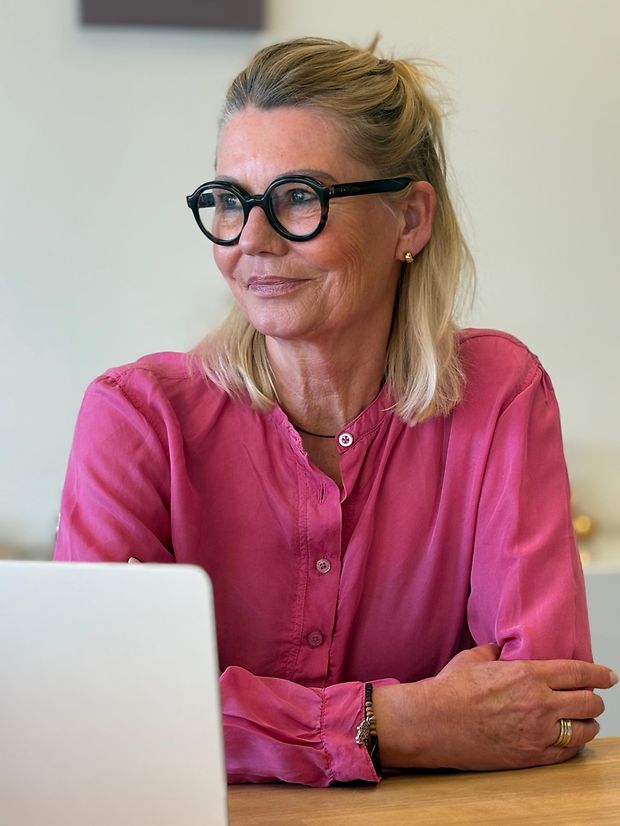 Eine Frau mit Brille und rosa Bluse sitzt und schaut zur Seite. 