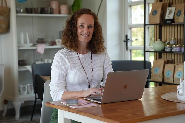 Eine Frau sitzt in einem Caf&eacute; an einem Laptop und schaut in die Kamera.