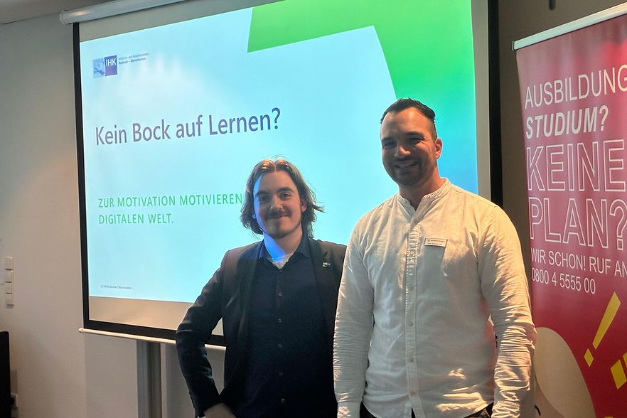 Zwei Personen stehen vor der Vortragspr&auml;sentation und l&auml;cheln in die Kamera.