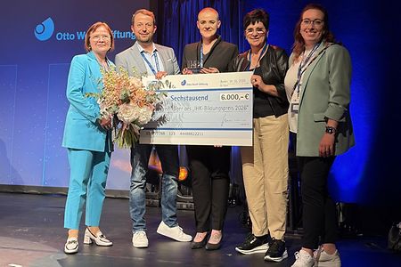 F&uuml;nf Personen auf einer B&uuml;hne halten einen gro&szlig;en Scheck &uuml;ber 6.000 Euro und einen Blumenstrau&szlig; bei einer Preisverleihung.