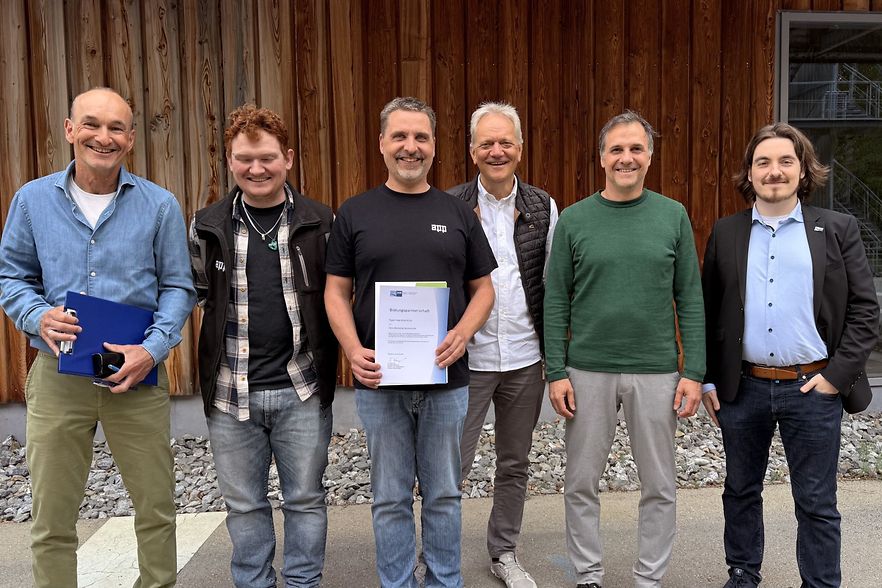 Sechs M&auml;nner stehen vor dem Schulgeb&auml;ude und l&auml;cheln in die Kamera.