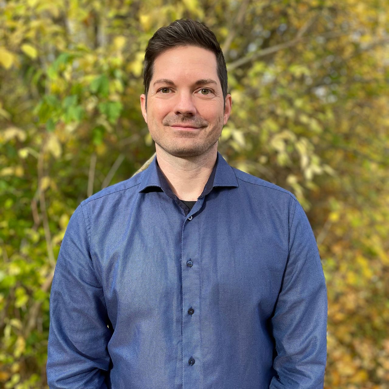 Porträt einer Person in blauem Hemd vor unscharfem Hintergrund mit herbstlich gefärbten Bäumen.