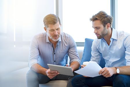 Zwei Businessm&auml;nner sitzen vor einem Tablet und f&uuml;hren ein Gespr&auml;ch.
