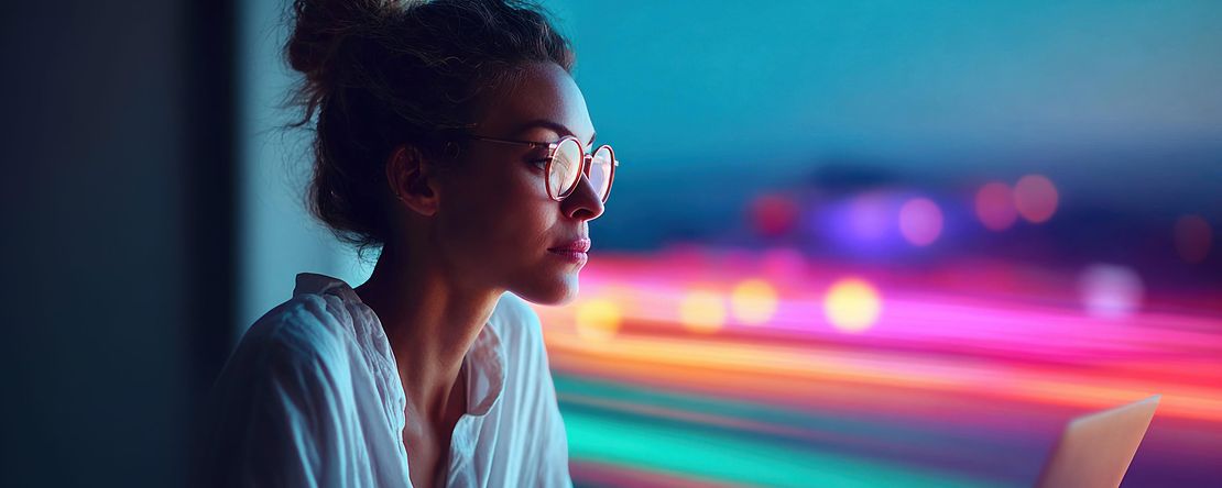 eine junge Frau arbeitet am Laptop (abends mit verzerrten Abendlichtern im Hintergrund)