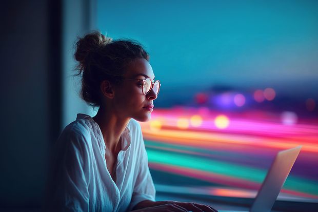 eine junge Frau arbeitet am Laptop (abends mit verzerrten Abendlichtern im Hintergrund)