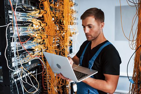 Junger Mann in Uniform arbeitet mit Laptop, Internetger&auml;ten und Kabeln im Serverraum