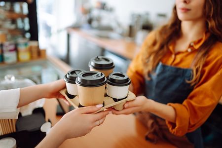 An einer Ladentheke werden vier Kaffee-to-go-Becher an eine Kundin &uuml;berreicht.