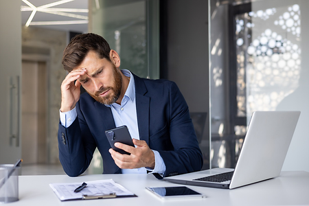 Ein Mann in Businesskleidung sitzt am Schreibtisch und schaut stirnrunzelnd auf sein Smartphone.