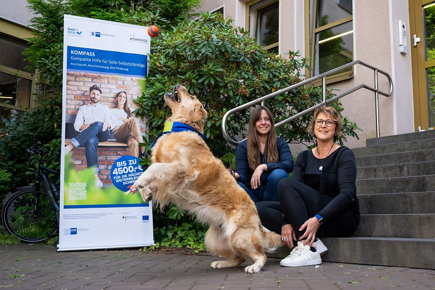 Zwei Frauen sitzen neben einem Roll-up auf einer Treppe. Im Vordergrund f&auml;ngt ein Hund einen Ball.