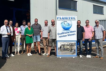 Eine Gruppe Menschen steht vor einem grauen Geb&auml;ude. In der Mitte steht ein blau-wei&szlig;es Rollup.